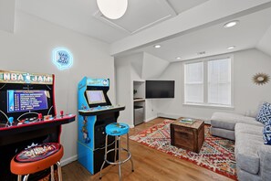 A living room with a basketball arcade game, a flat-screen TV, a sofa, and a coffee table. - Escape to this spacious three-level home in the heart of Ocean Grove, just a short walk from the beach, boardwalk, shops, and restaurants. (Neptune Township)