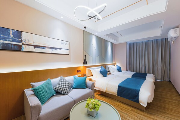 A modern hotel room with a sofa, two beds, a coffee table with a flower arrangement, a painting on the wall, and a ceiling light fixture.
