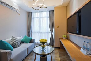 A modern hotel room with a sofa, a small round table with a vase of flowers, a flat-screen TV mounted on the wall, and a water dispenser on the sideboard.