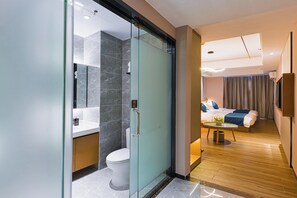 A modern hotel room with a glass partition separating the bathroom, a wooden floor, a bed with a blue blanket, and a small table with a plant.