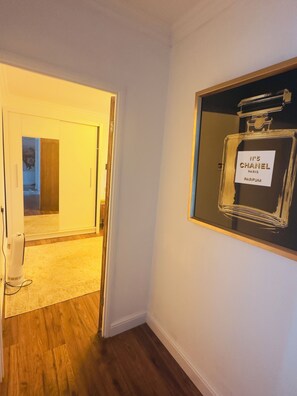 Un couloir avec un plancher en bois, une porte donnant sur une autre pièce et un flacon de parfum encadré accroché au mur.