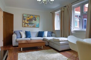 A living room with a sectional sofa, a coffee table, a painting on the wall, and a window with curtains. - Elegant Wimbledon Apartment (London)