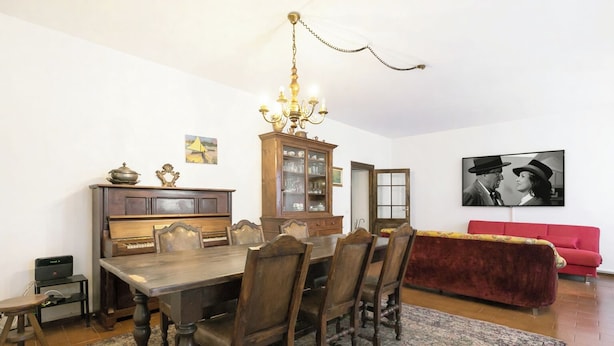 A dining area with a wooden table and chairs, a chandelier, a cabinet with glassware, and a red sofa.