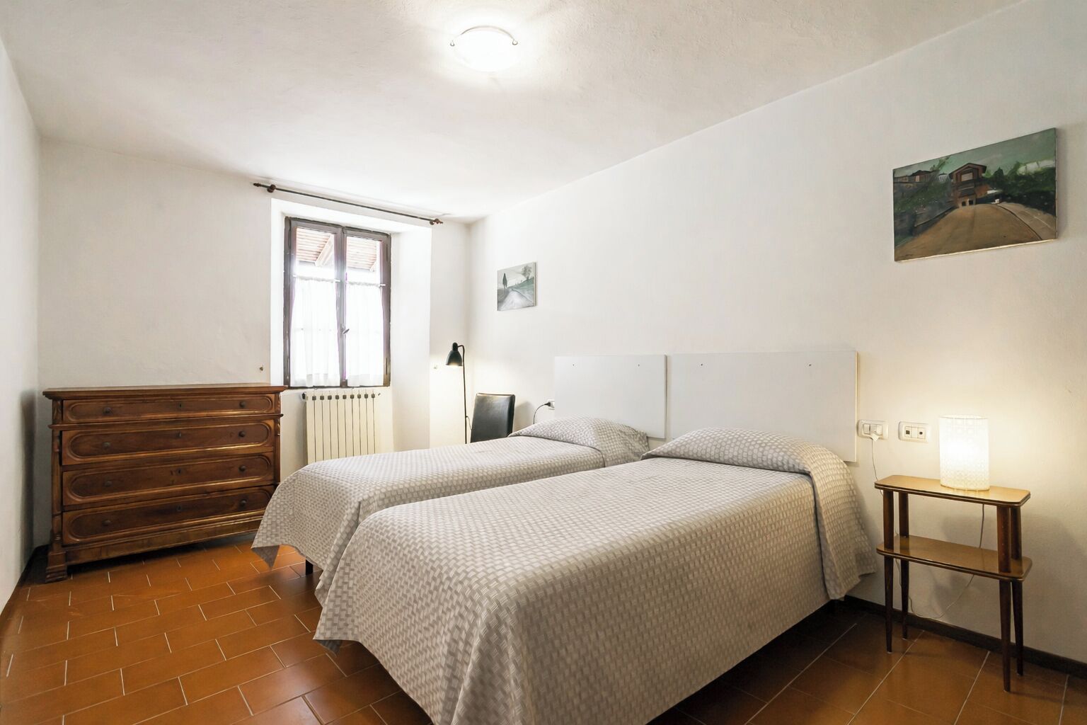 A bedroom with two beds, a dresser, a radiator, a window, and a painting on the wall.