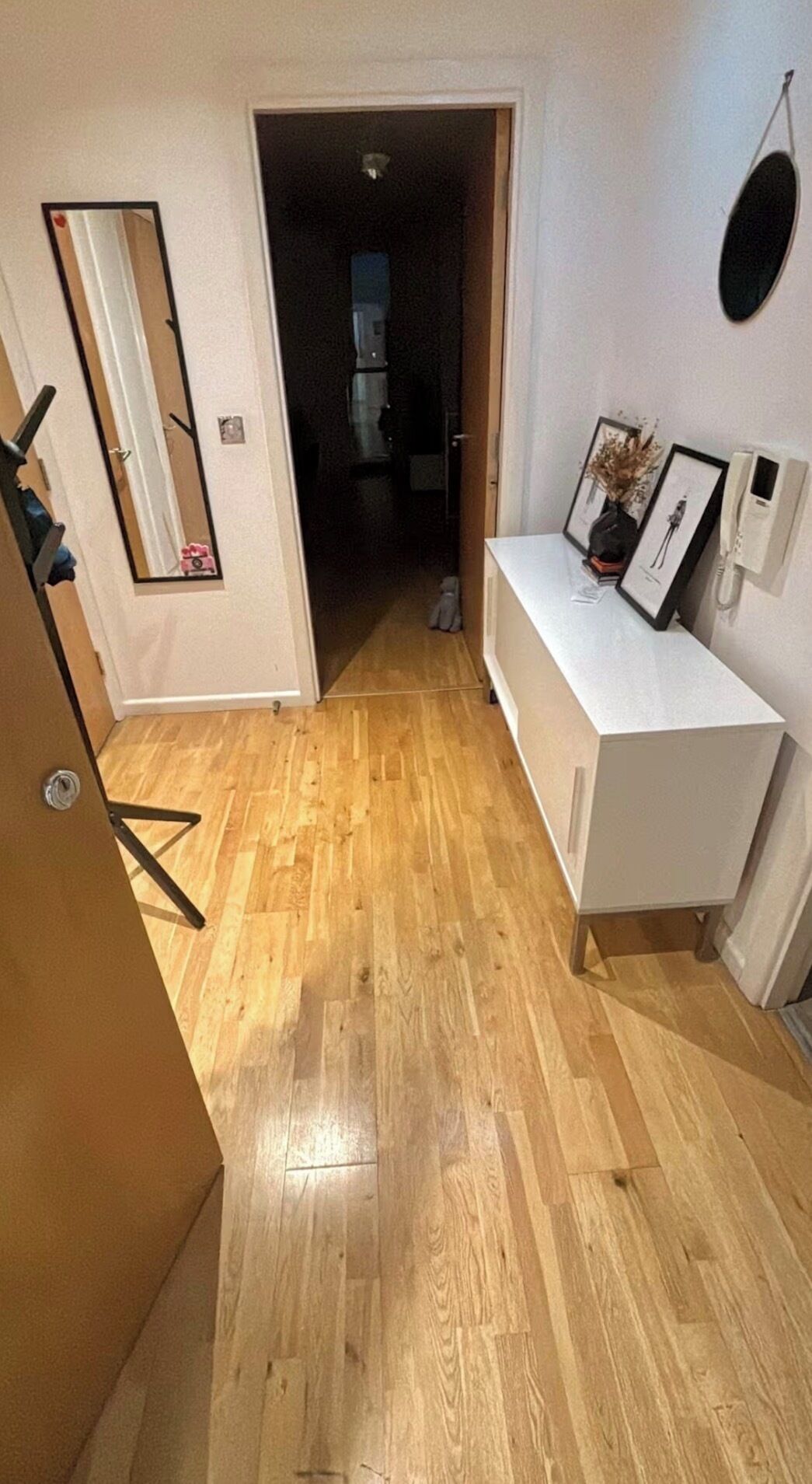 A hallway with a wooden floor, a white console table with framed pictures, a mirror, and a door leading to another room.