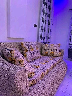 A woven sofa with patterned cushions in a room with a tiled floor and a wall-mounted light fixture.