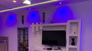 Una sala de estar moderna con un televisor de pantalla plana montado en un mueble blanco, un sofá amarillo y una mesa de comedor con mantel estampado.