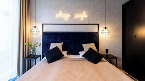 Quarto moderno com cabeceira escura, um letreiro de neon com a inscrição 'stay safe' e duas luminárias de mesa.