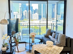 Uma sala de estar moderna com um sofá, uma mesa de centro e uma televisão. Há uma varanda com vista para a cidade.