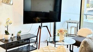 A modern living room with a flat-screen TV on a tripod stand, a glass coffee table, a sofa, and a small side table with a lamp. - Prime 2BR Location - Pool & Parking @BCT 1004 (South Brisbane)