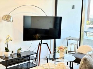 Uma sala de estar moderna com uma televisão de tela plana em um suporte de tripé, uma mesa de centro de vidro, um sofá e uma pequena mesa lateral com um abajur.