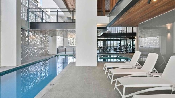 A modern indoor swimming pool with lounge chairs and a view of a gym area. - Prime 2BR Location - Pool & Parking @BCT 1004 (South Brisbane)