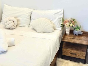 Uma cama com um edredom branco e almofadas, uma mesa de cabeceira de madeira com um vaso de flores e um tapete branco fofo.