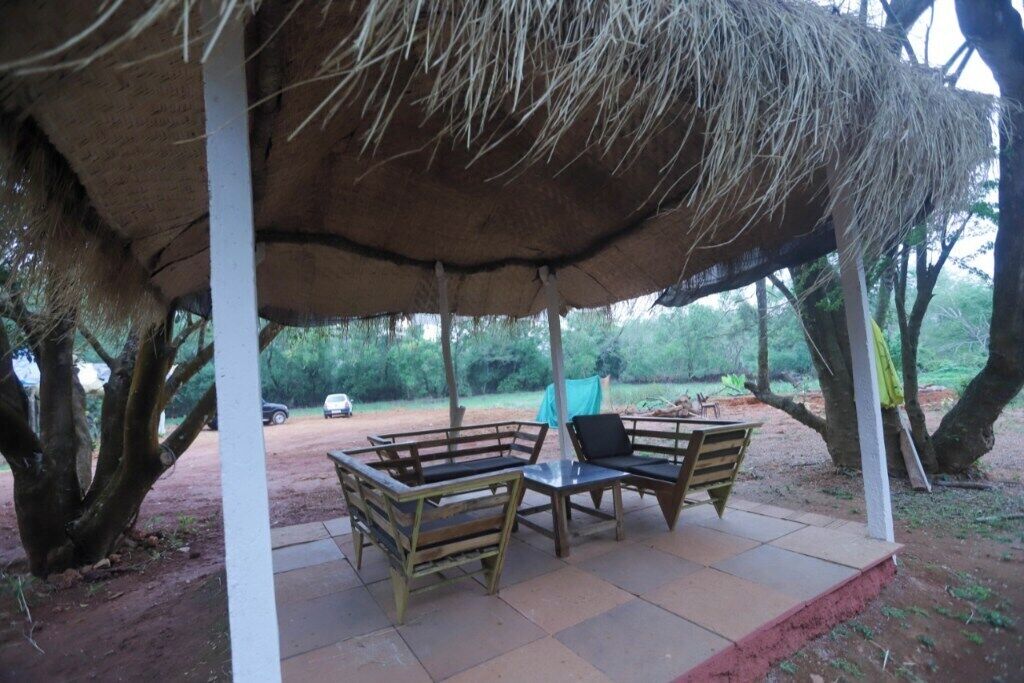 Un espacio de estar techado al aire libre con bancos y una mesa, rodeado de árboles y un sendero de tierra.