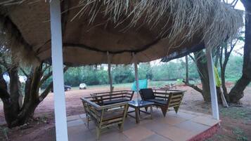 Un espacio de estar techado al aire libre con bancos y una mesa, rodeado de árboles y un sendero de tierra.