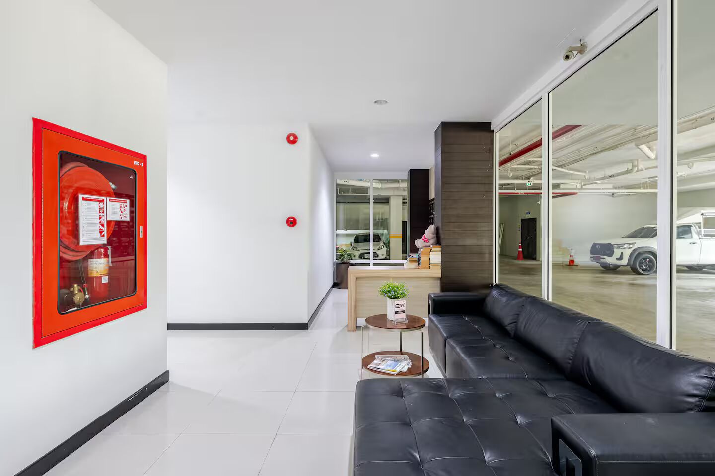 Un interior moderno con un sofá de cuero negro, un detector de incendios rojo y vistas a un estacionamiento a través de amplias ventanas.