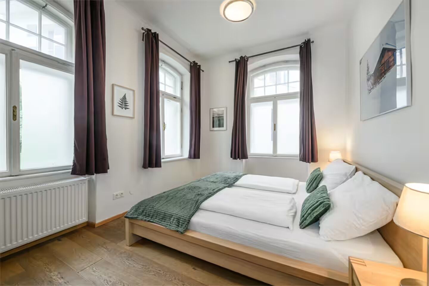 Un dormitorio con una cama, dos ventanas, cortinas, un radiador y un cuadro en la pared.