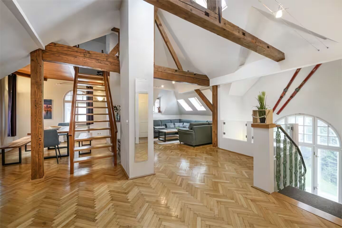 A spacious loft with high ceilings, exposed wooden beams, a herringbone wooden floor, a staircase, a seating area with a sofa and table, and large windows.