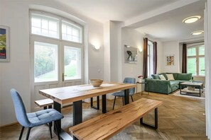 Un espace repas comprenant une table et des chaises en bois, un banc et une vue sur un espace extérieur verdoyant, visible à travers de grandes fenêtres.