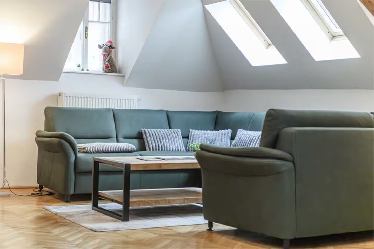 Ein gemütliches Wohnzimmer mit einem grünen Sofa, einem Holztisch und einem einzelnen Sessel. Über dem Sofa befinden sich Dachfenster und darunter ein Heizkörper.
