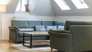 Un salon chaleureux avec un canapé vert, une table basse en bois et un fauteuil. Des fenêtres de toit sont situées au-dessus du canapé et un radiateur en dessous.