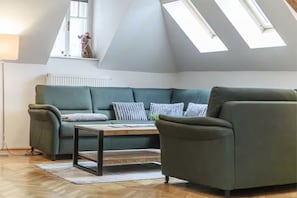 Ein gemütliches Wohnzimmer mit einem grünen Sofa, einem Holztisch und einem einzelnen Sessel. Über dem Sofa befinden sich Dachfenster und darunter ein Heizkörper.