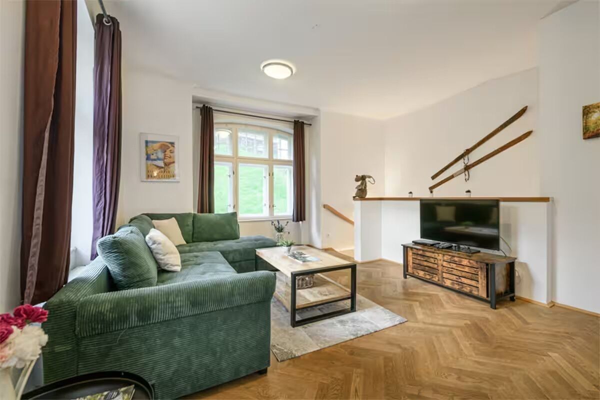 Una sala de estar con un sofá verde, una mesa de centro, un televisor sobre un mueble de madera y una ventana con cortinas.