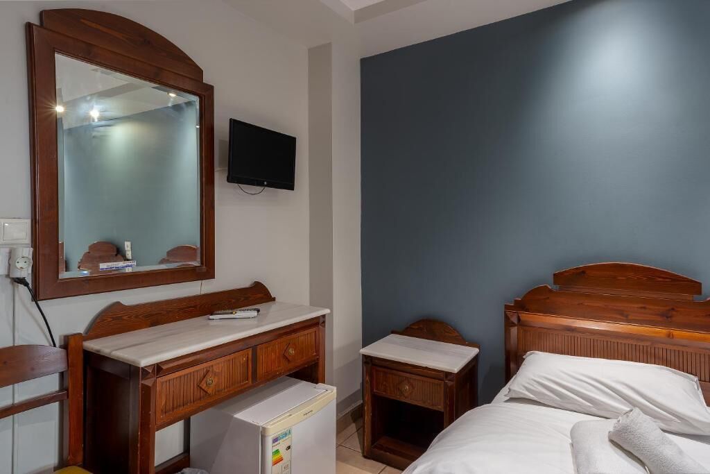 A bedroom with a wooden bed, a small bedside table, a mirror, a television mounted on the wall, and a refrigerator.