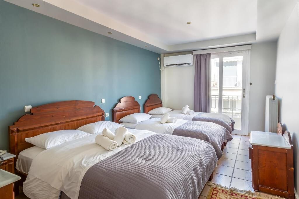 Ein Hotelzimmer mit zwei Betten, einem Holz-Kopfteil, einem kleinen Nachttisch, einer Fensterbank und Vorhängen sowie einer Klimaanlage.