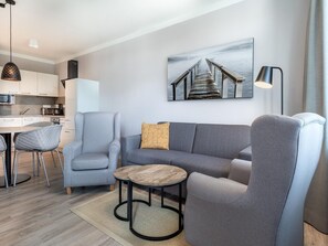 Ein modernes Wohnzimmer mit einem grauen Sofa, einem kleinen runden Tisch und einem Sessel. Es gibt eine Küchenzeile mit weißen Schränken und einer Mikrowelle. An der Wand hängt ein Gemälde einer Brücke.