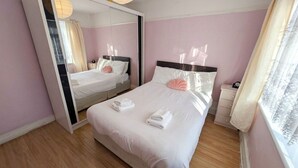 A hotel room with a large bed, a bedside table, a mirror, and a window with curtains. - Peaceful 4BDR Home with Garden in Slough (Berkshire)