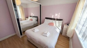 A hotel room with a large bed, a bedside table, a mirror, and a window with curtains. - Peaceful 4BDR Home with Garden in Slough (Berkshire)