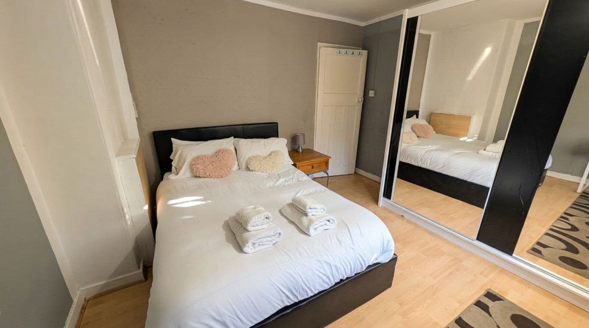 A neatly made bed with white linens and two folded towels, a bedside table with a lamp, and a large mirror reflecting the room.