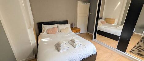 A neatly made bed with white linens and two folded towels, a bedside table with a lamp, and a large mirror reflecting the room.