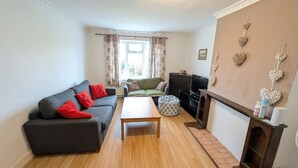 A living room with a sofa, ottoman, and a coffee table. There is a fireplace with decorative items and a television. - Peaceful 4BDR Home with Garden in Slough (Berkshire)