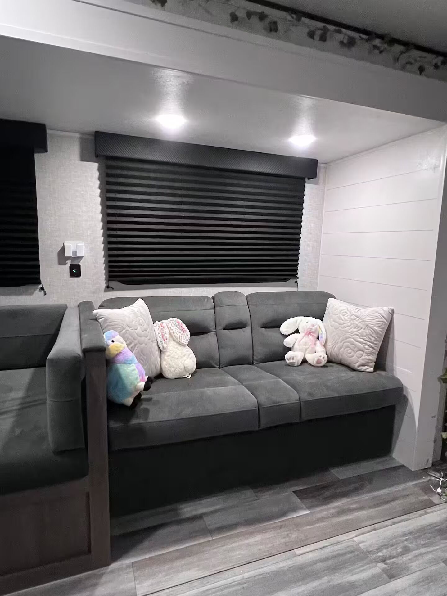 Un sofá gris con una banqueta integrada, una ventana con persianas y una lámpara fijada a la pared.