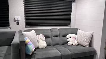 Um sofá cinzento com um banco embutido, uma janela com persianas e uma luminária fixada na parede.
