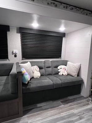 En grå sofa med indbygget bænk, et vindue med rullegardiner og en vægmonteret lampe.
