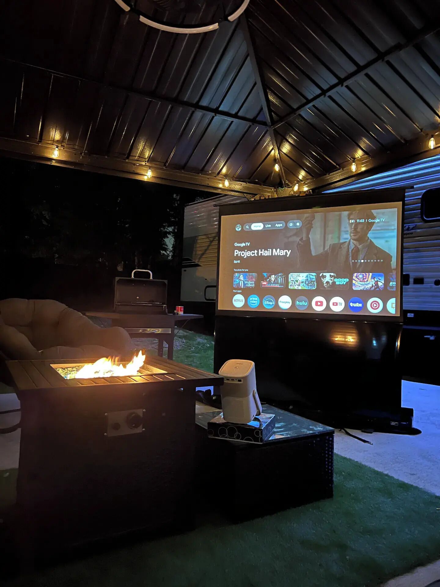 Un área de estar al aire libre acogedora con un brasero, una pantalla de proyector mostrando una serie de televisión y una parrilla.