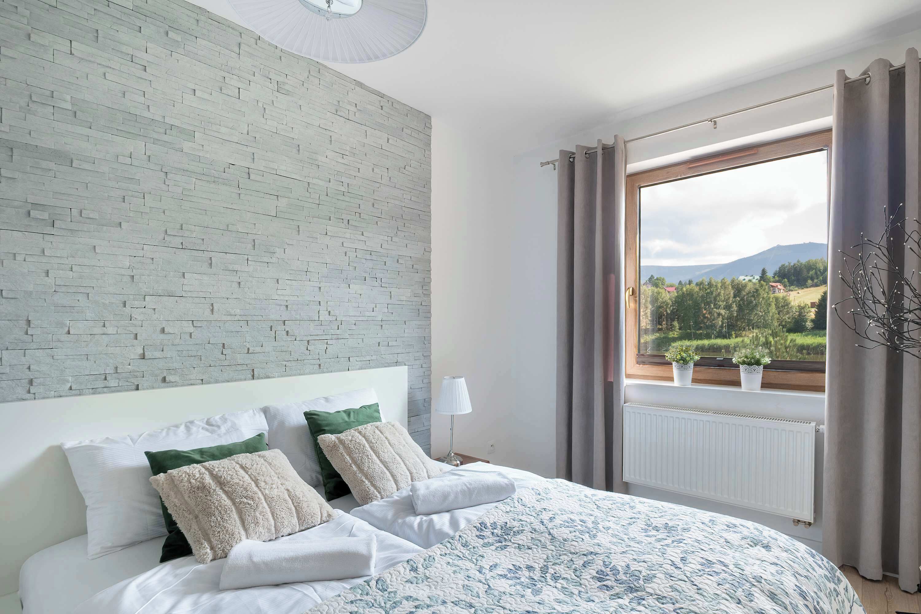 Un dormitorio con pared de ladrillo, una cama grande con muchas almohadas, una ventana con cortinas y vista a un paisaje.