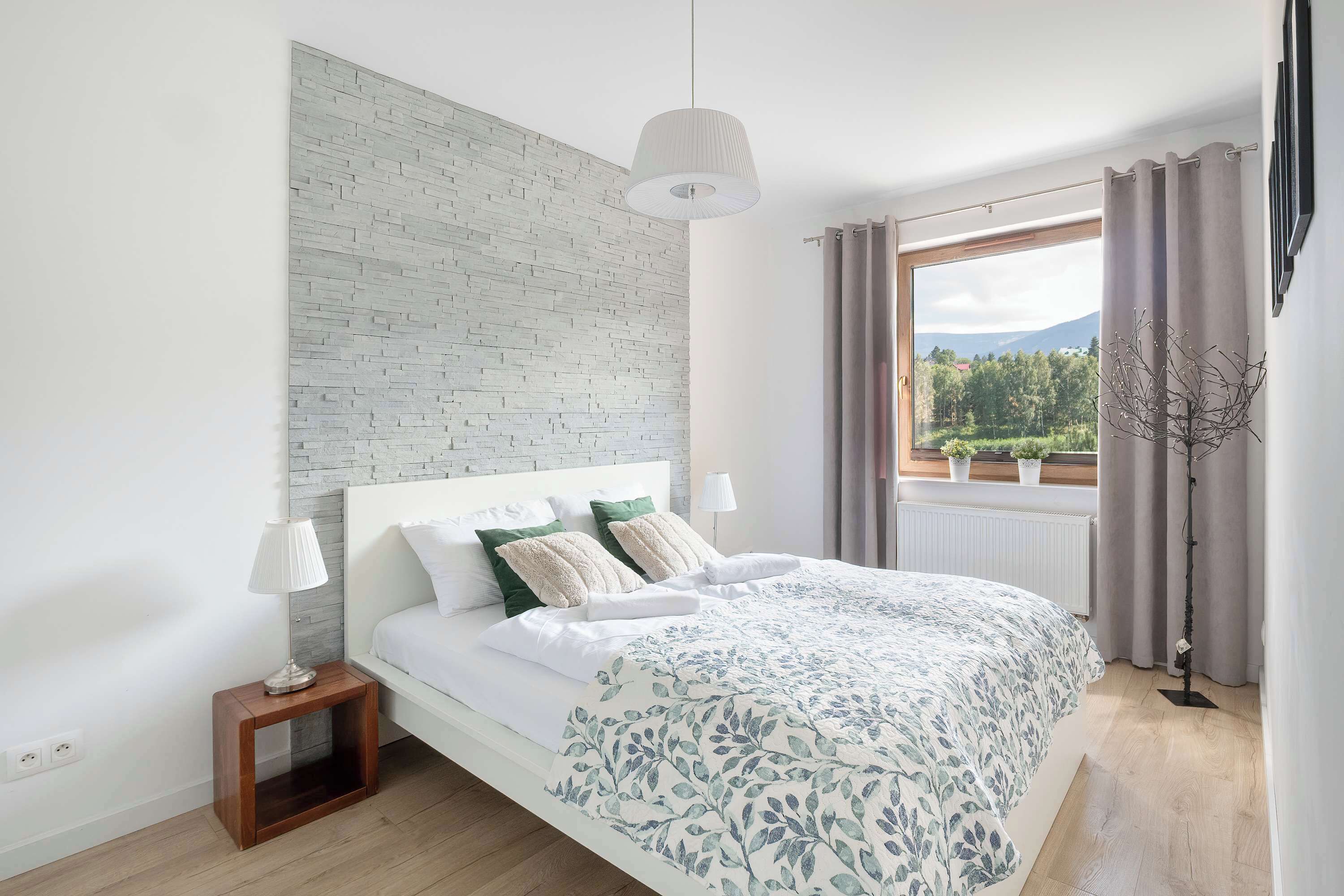 Une chambre à coucher avec un lit, des lampes de chevet, une table de nuit, une fenêtre avec des rideaux et une vue sur la verdure à l’extérieur.