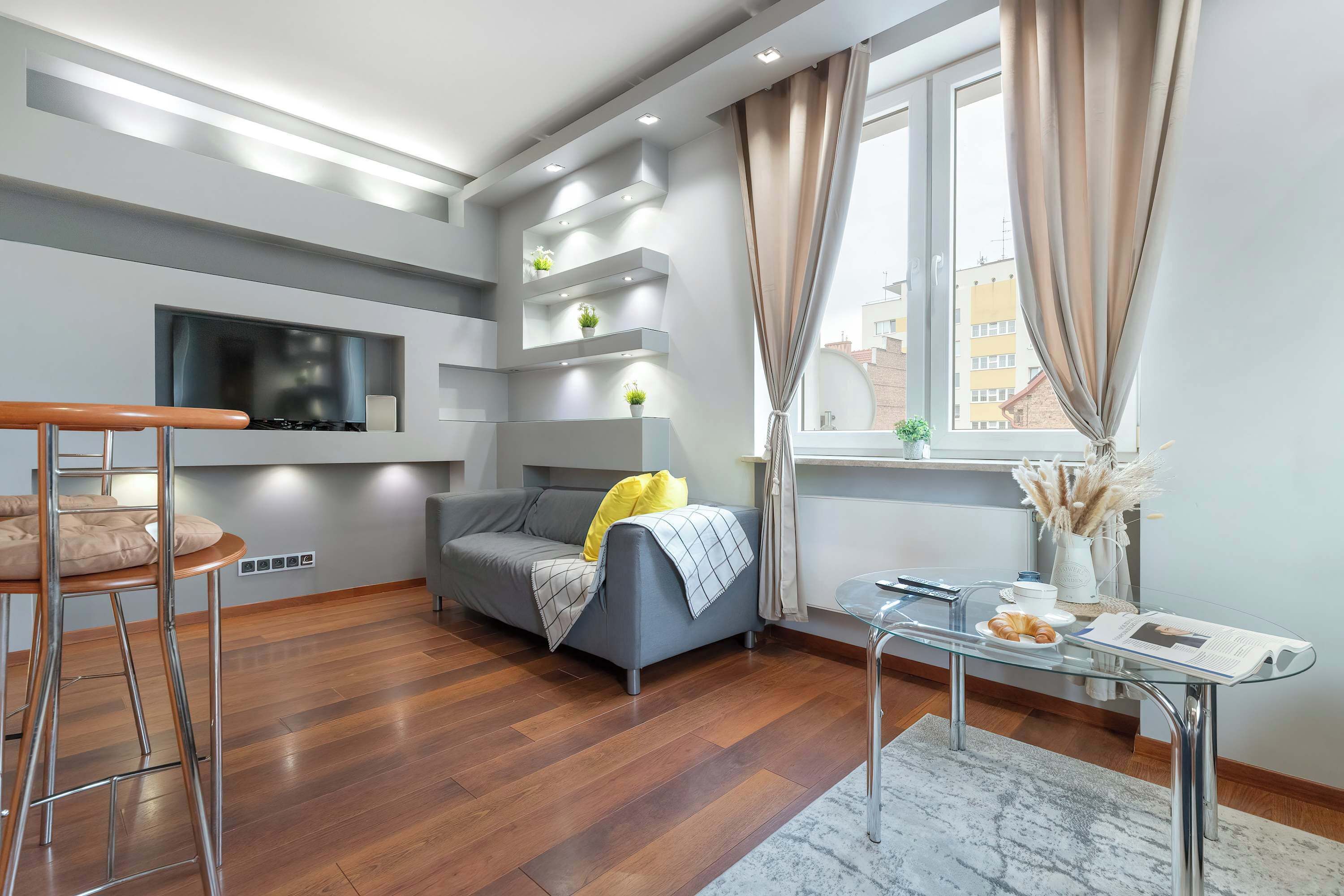 Una sala moderna con un sofá gris, una mesa de comedor de madera y un televisor montado en la pared.