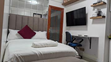 Un dormitorio con cama, un banco, un escritorio con silla, un televisor y una puerta.