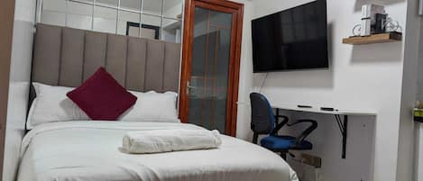 A bedroom with a bed, a bench, a desk with a chair, a television, and a door.