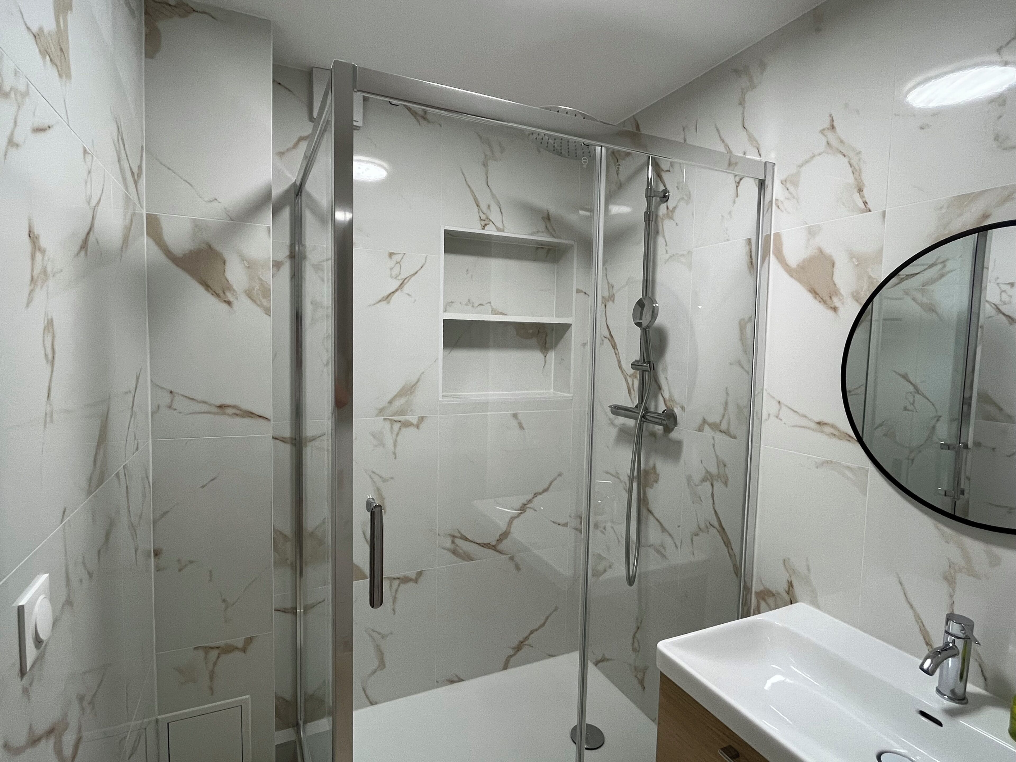 Une salle de bain moderne avec une douche à parois de verre, un lavabo blanc et un mur décoré d’un motif imitant le marbre.