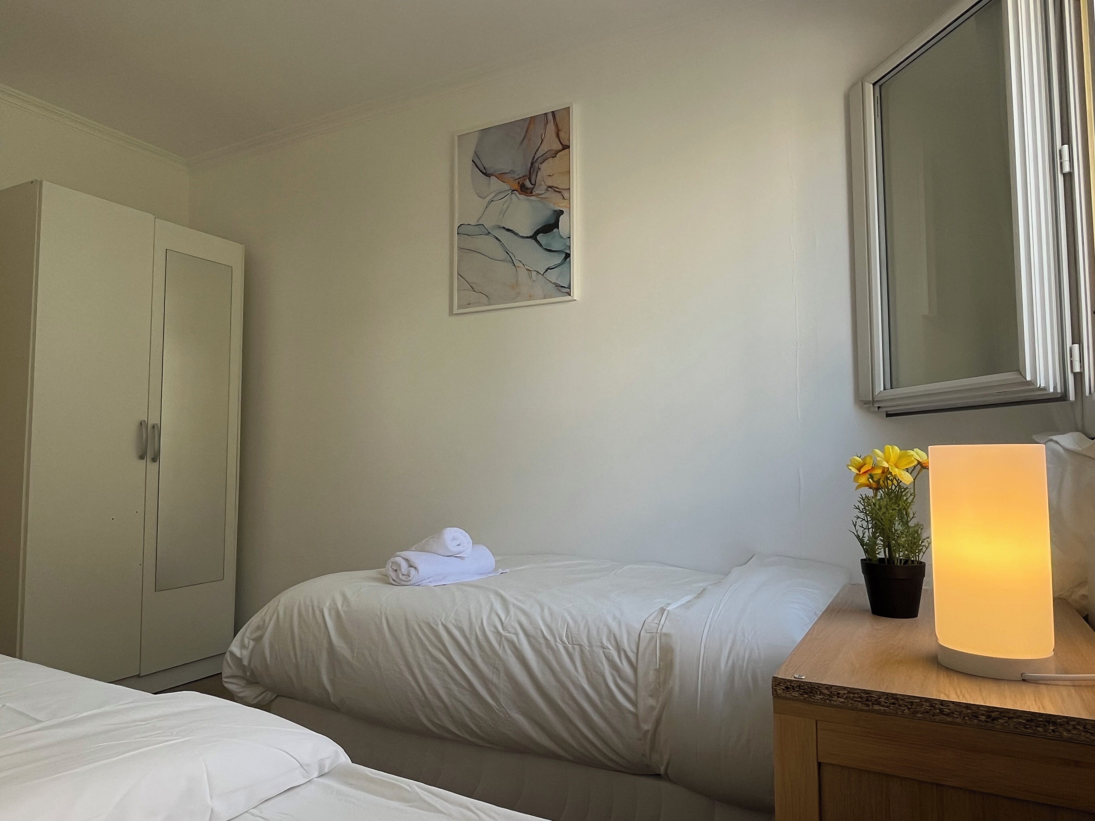Ein Hotelzimmer mit Bett, Schrank, einem Gemälde, einer Topfpflanze und einer Nachttischlampe.