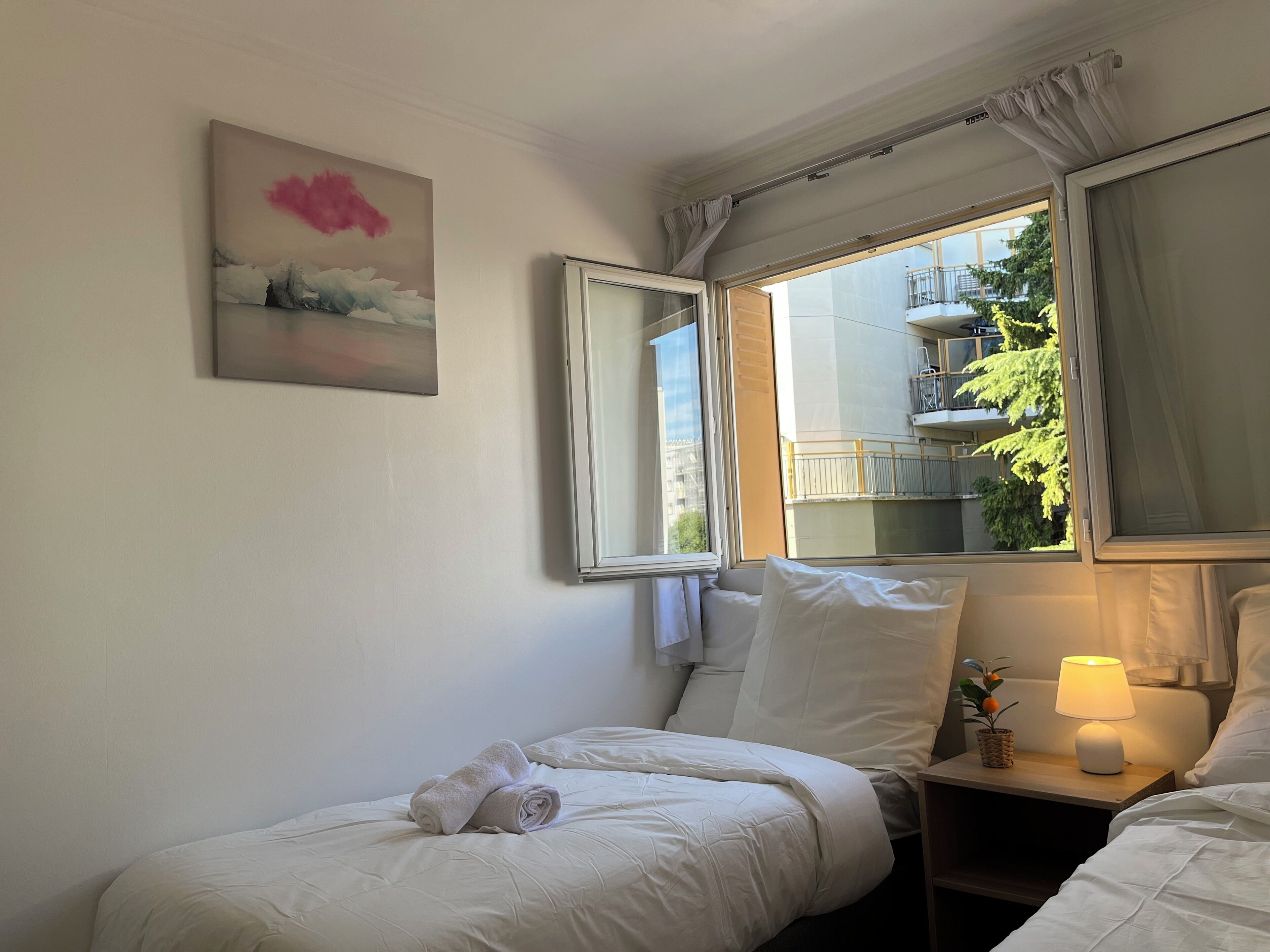 A hotel room with a bed, a bedside table with a lamp, a window with curtains, and a painting on the wall.
