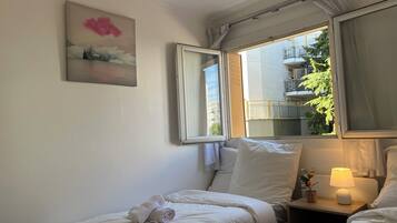 A hotel room with a bed, a bedside table with a lamp, a window with curtains, and a painting on the wall.