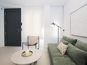 Een minimalistische woonkamer met een groene bank, een ronde salontafel met een kaars, een zwarte stoel, een vloerlamp en een deur.
