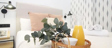 A neatly made bed with a white headboard, a single pink pillow, and a woven basket containing a glass of orange juice, a white bowl, and a white cup.
